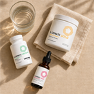 Lumati Gold powder tub, Lumati Green 100 mg capsule bottle, and Lumati Red dropper bottle on a beige linen napkin beside a glass of water in warm sunlight.